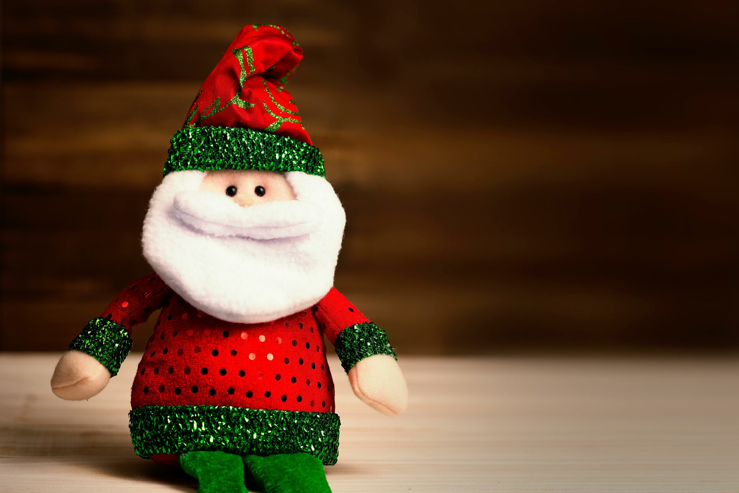 Cute Santa Claus plush toy wearing red and green attire sits on a wooden surface, perfect for holiday decor.