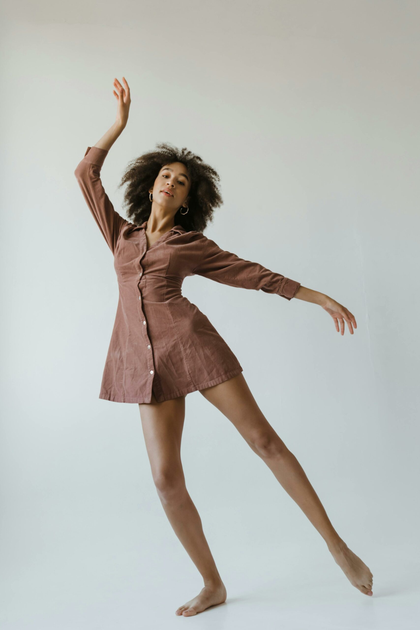 Elegant young woman with curly hair dancing gracefully in a brown dress, embodying beauty and empowerment.